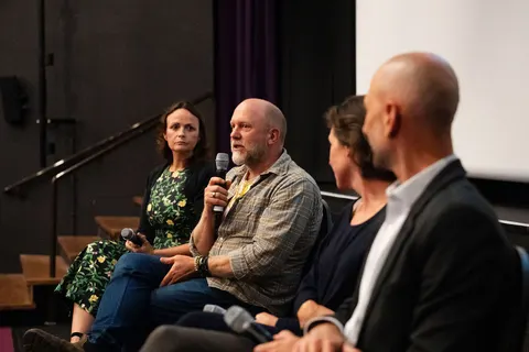 Audience members seated close to the camera, one holding a microphone to speak