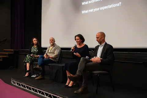 Four people seated on chairs on the stage for the panel discussion, a question projected behind them