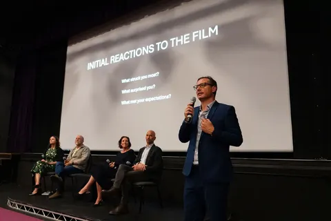 A man standing with a microphone in front of the screen, with panellists seated behind him