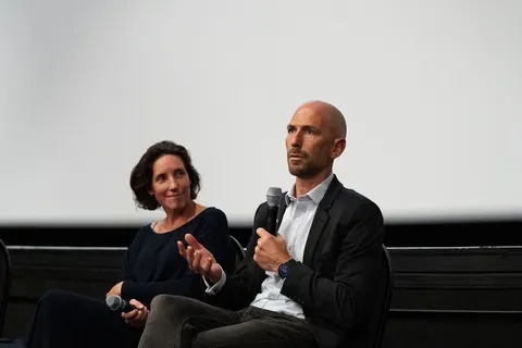 Two panellists seated side by side, a woman and a man holding a microphone, in front of a white screen