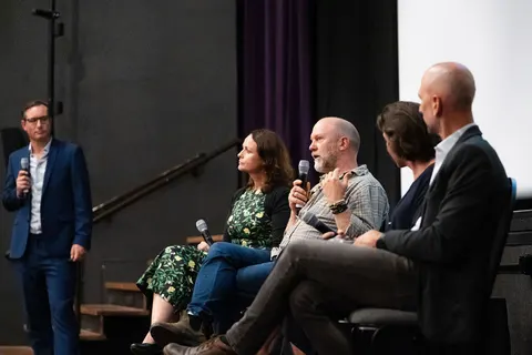 A seated bearded panellist speaking into a microphone, with a standing facilitator at the left of the stage