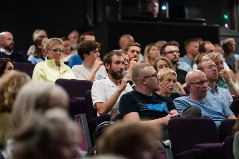 Audience members seated in a row, some looking toward the stage