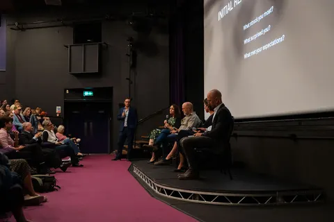 Two speakers seated on the stage beside the screen while the audience looks on